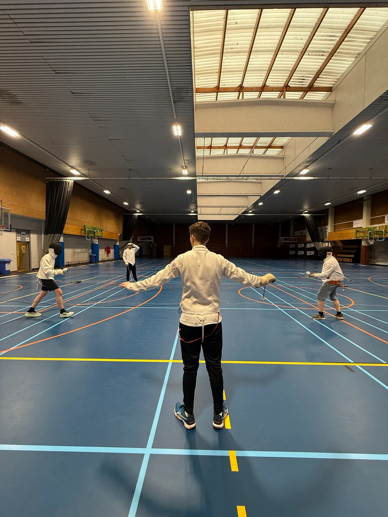 Een trainer staat met gespreide armen midden op de vloer terwijl de deelnemers achter hem per twee schermen