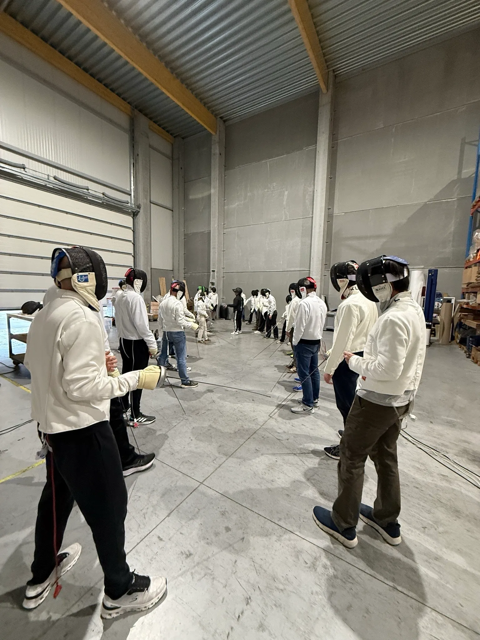 Deelnemers in schermvest en masker staan per twee tegenover elkaar in een bedrijfshal