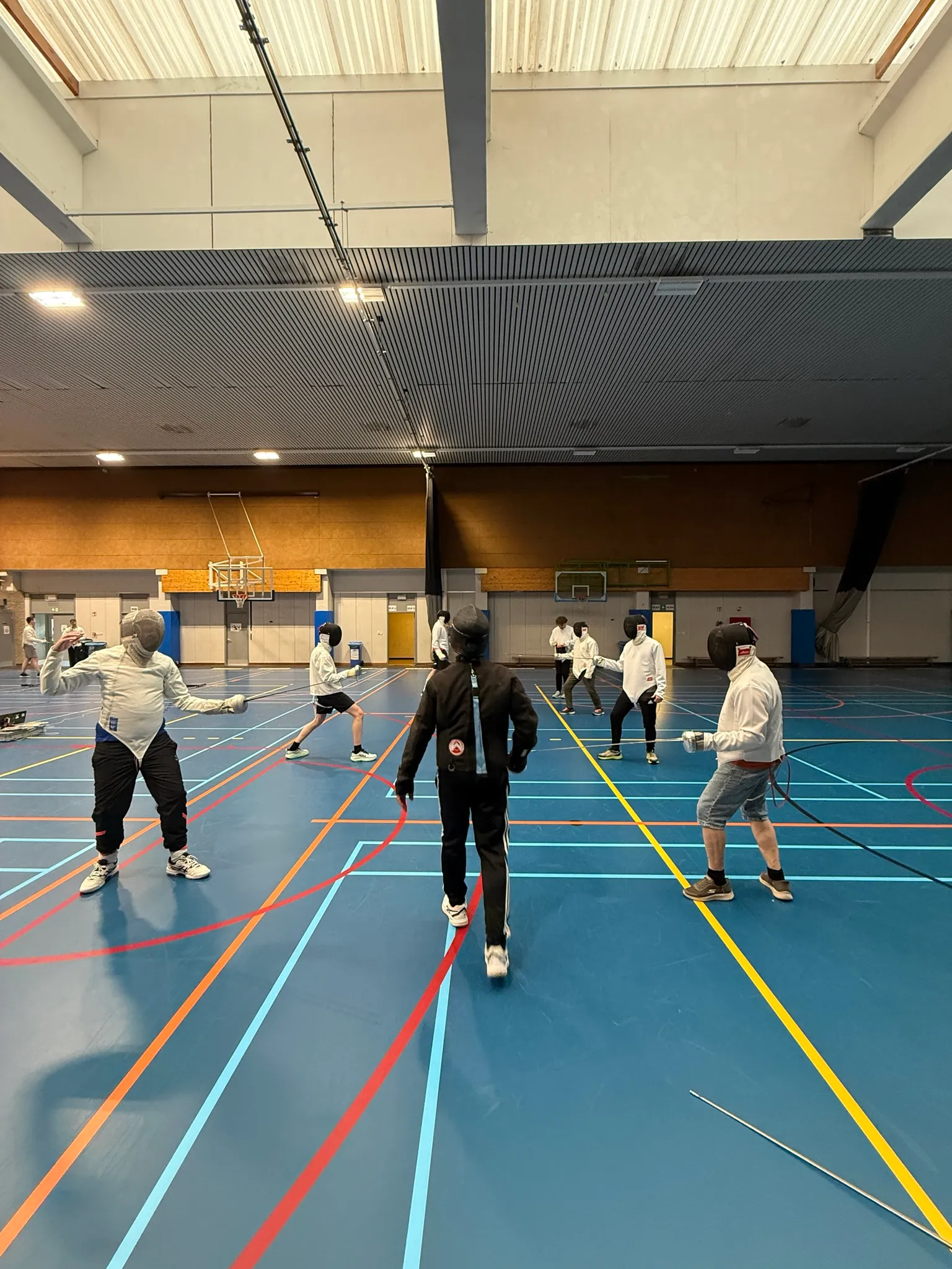 Deelnemers schermen per twee verspreid over een sporthal, terwijl een trainer middenin toekijkt